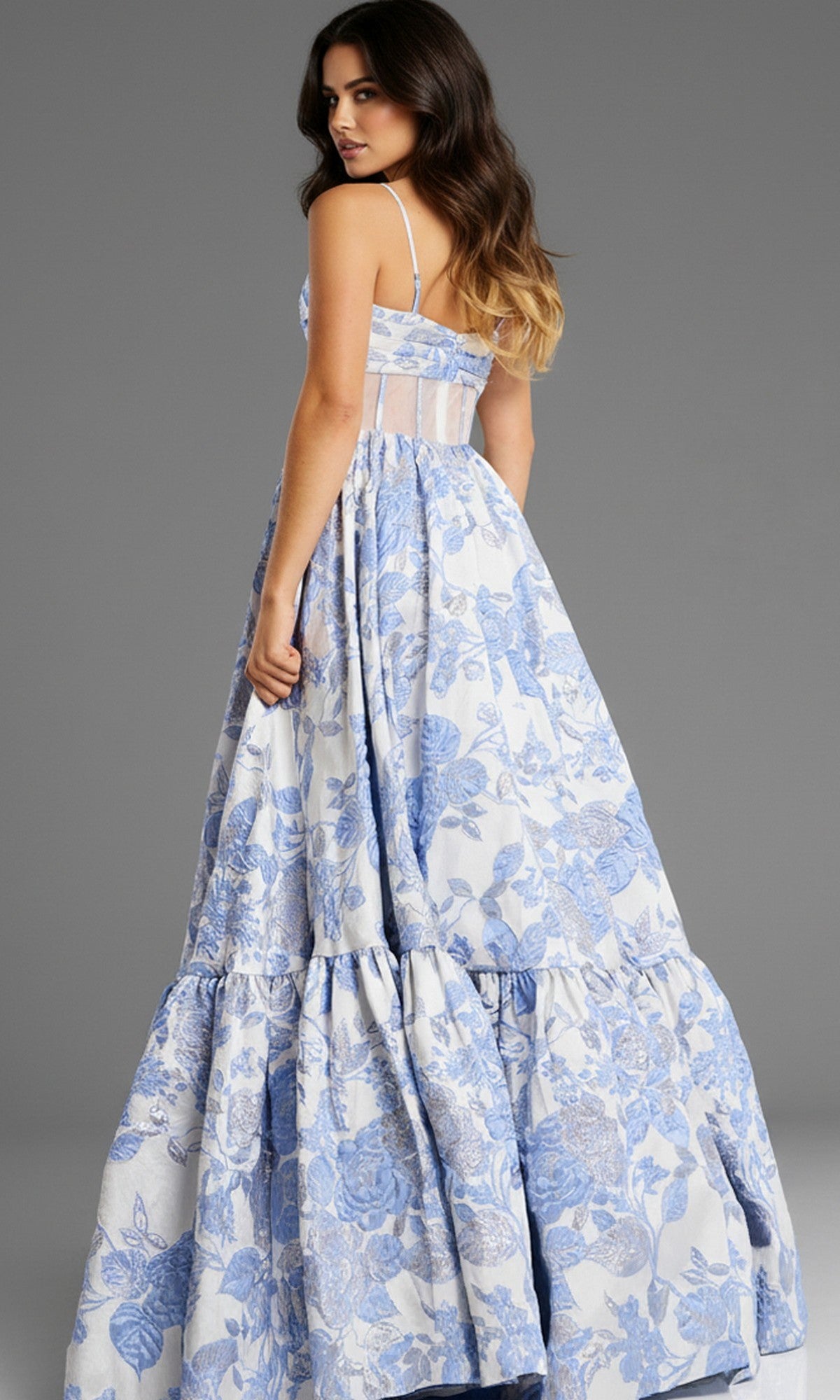 A woman in the Jovani 47328 long prom dress—sleeveless, with thin straps, a blue and white floral jacquard pattern, and an elegant A-line skirt—stands looking over her shoulder against a plain gray background.